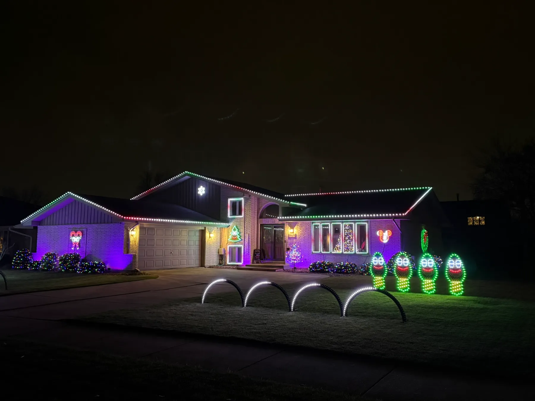 The front of the house lit purple, with white arches on the lawn and the four singing bulbs glowing green.