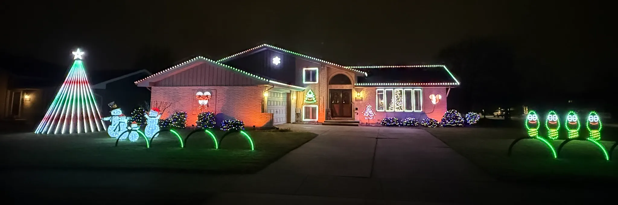 Our house at night last November, seen from across the street, with each part of the show labeled: the mega tree and its star on the left, the rooflines, windows, snowflakes, candy canes and spinner on the house, the bushes and floodlights below them, arches on both sides of the driveway and the four singing bulbs on the right.